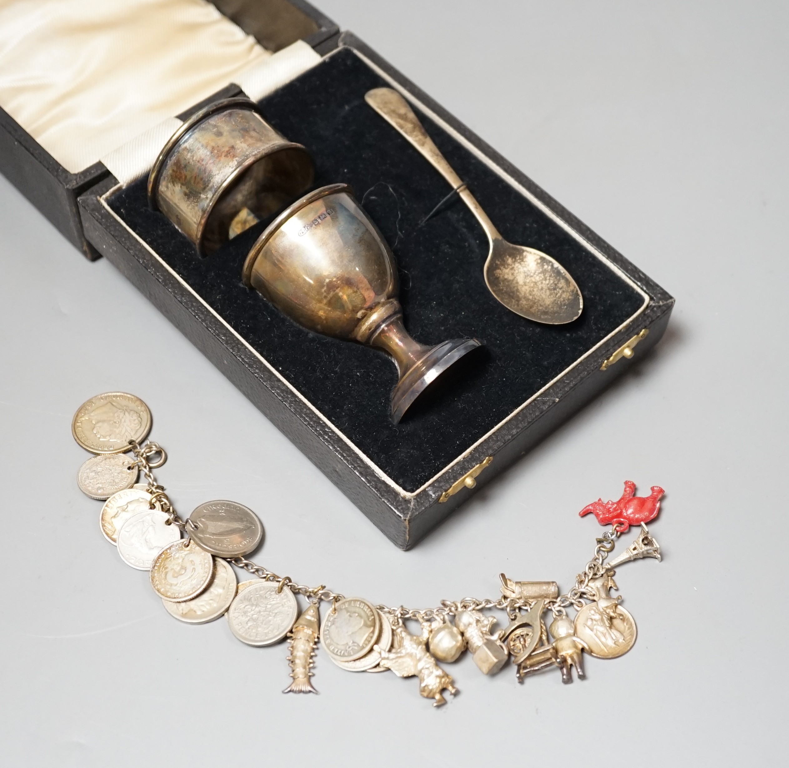 A 1930's cased silver christening set, comprising an egg cup, spoon and napkin ring, together with a white metal charm bracelet hung with assorted charms and coins including silver.